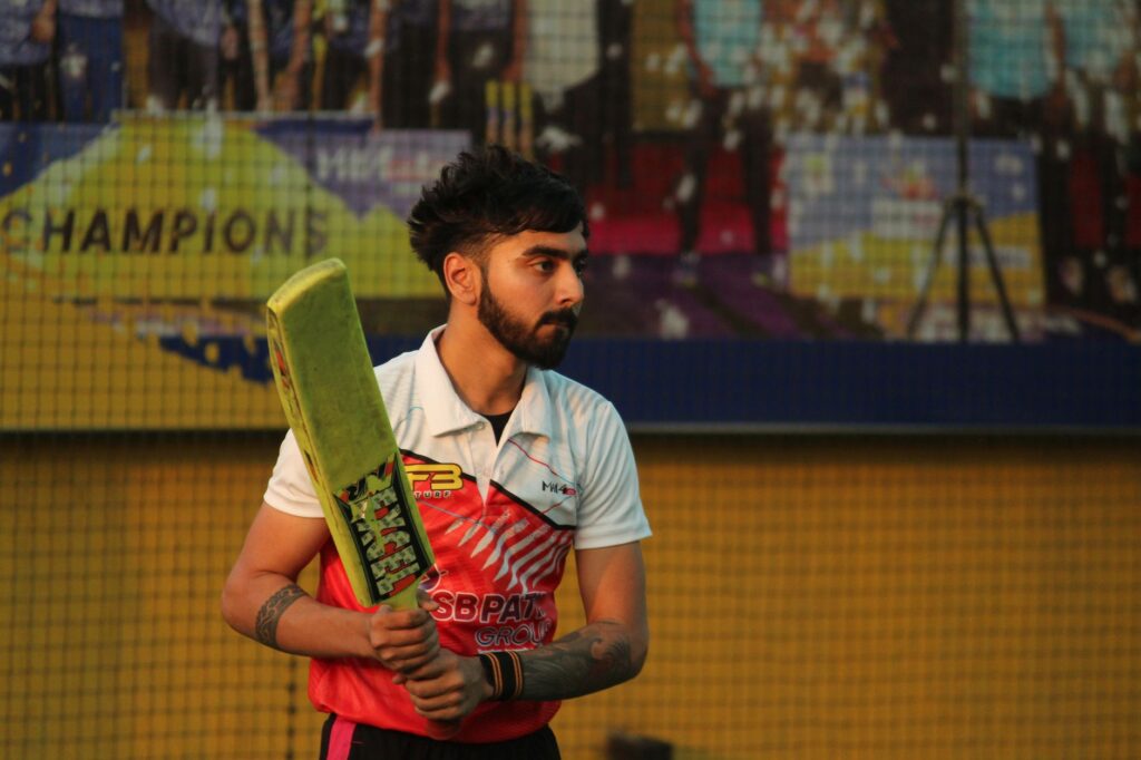 A focused cricketer holding a bat, ready to play on an artificial turf field. The backdrop includes a colorful advertisement and a netted area.