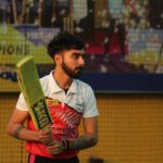A focused cricketer holding a bat, ready to play on an artificial turf field. The backdrop includes a colorful advertisement and a netted area.