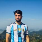 A man in an Argentina football jersey stands with a scenic mountain backdrop under a clear sky.