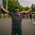 Joyful soccer fan with arms raised, celebrating outdoors in casual sportswear.