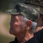 Elderly Asian man wearing a camouflage military cap, outdoors.