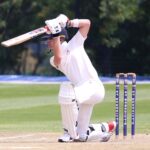 Cricketer in action performing a sweep shot on a sunny outdoor pitch.