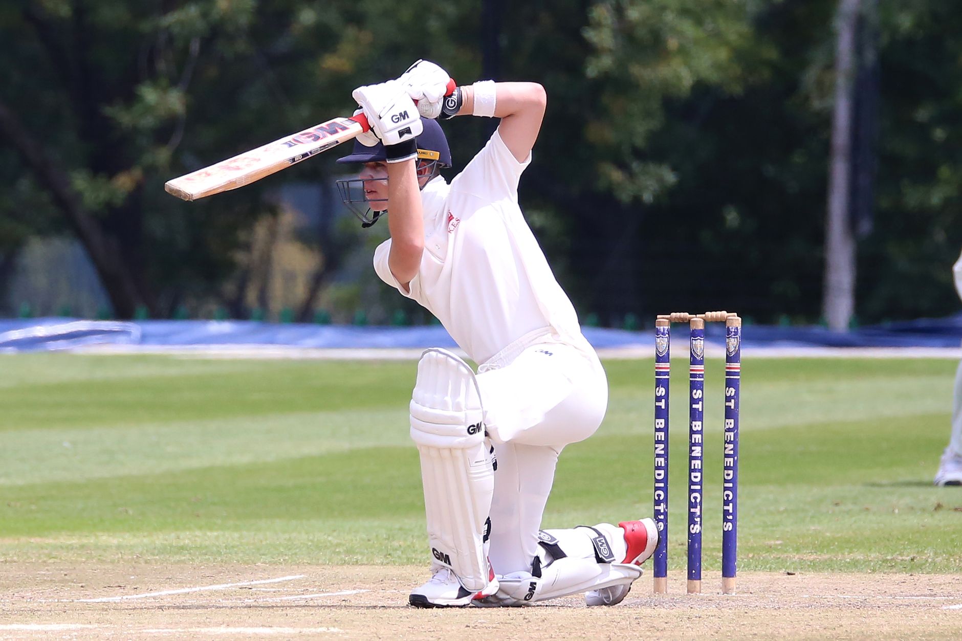 Cricketer in action performing a sweep shot on a sunny outdoor pitch.