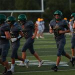 High school football team running on field during sunset, wearing green uniforms and helmets.