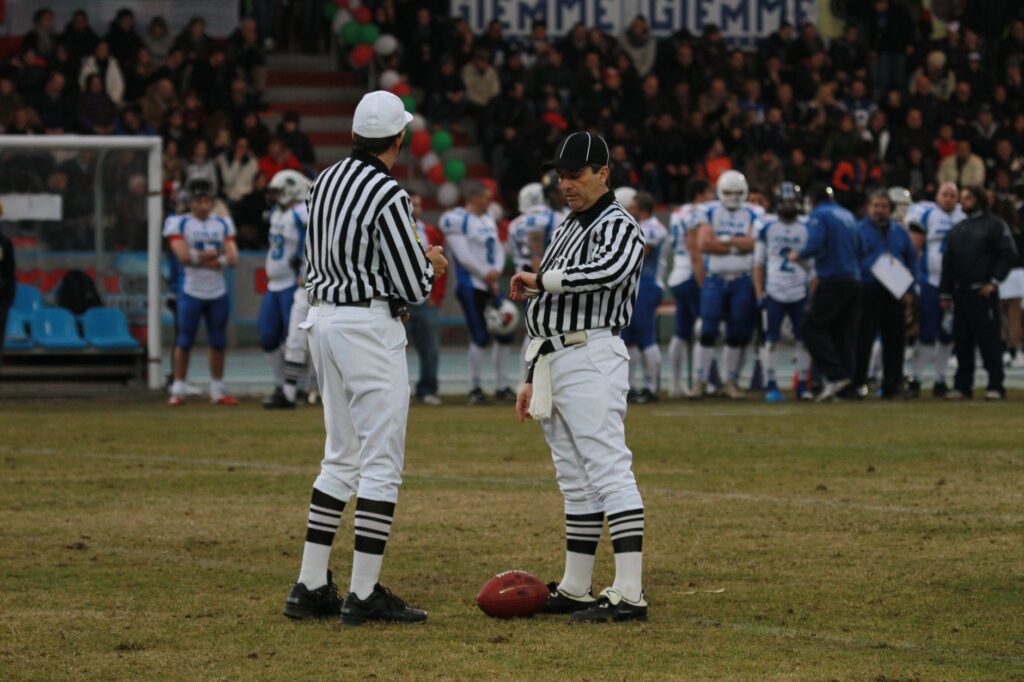 Two referees on a football field preparing for a game with players in the background at a stadium.