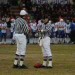 Two referees on a football field preparing for a game with players in the background at a stadium.