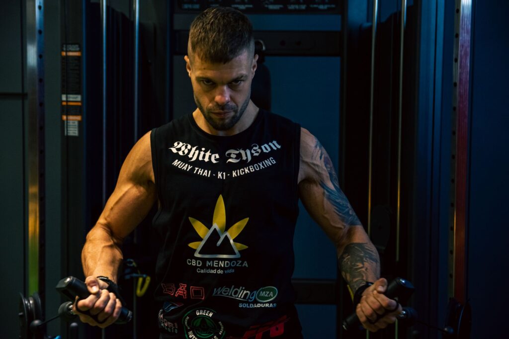 Focused man with tattoos works out in a dimly lit gym, showcasing strength and determination.
