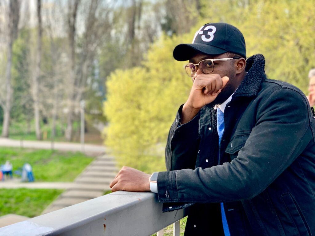 African American man in a black jacket and cap, posing thoughtfully on a balcony outdoors in spring.
