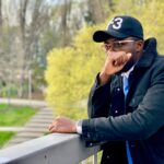 African American man in a black jacket and cap, posing thoughtfully on a balcony outdoors in spring.