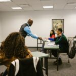 College classroom scene with diverse students and a teacher distributing papers, promoting education.