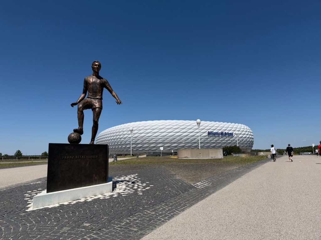 Statue of Franz Beckenbauer with Allianz Arena in Munich, Germany under a clear blue sky.