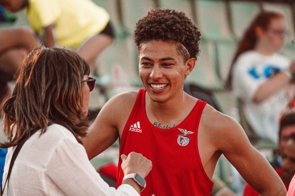 Young athlete in red jersey smiling during a post-event interaction with a woman at a sports venue.