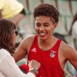 Young athlete in red jersey smiling during a post-event interaction with a woman at a sports venue.