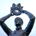 Bronze statue of a figure holding a crown aloft, located outdoors in Warwickshire, England.