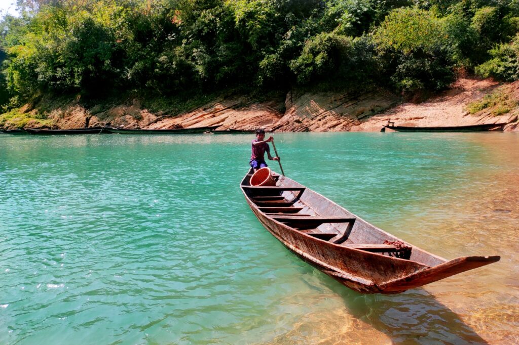 A person rowing a wooden boat on turquoise waters surrounded by lush greenery in Chattogram, Bangladesh.