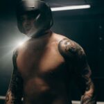 Focused boxer with tattoos training in dimly lit gym, wearing head guard.