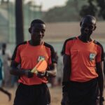 Two soccer referees in orange jerseys walking during a daytime match.