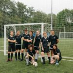 A diverse group of women soccer players pose confidently on a green soccer field.