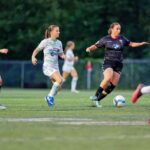 Dynamic action shot of female soccer players in motion during a game on a grass field.