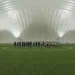Two soccer teams lined up on an indoor artificial turf field under dome lighting.