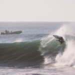 Surfer skillfully rides a powerful wave at Jeffreys Bay, South Africa, with support crew nearby.