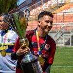 Two soccer players celebrate with a championship trophy on the field.