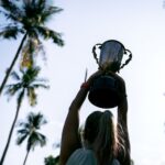 A woman lifts a trophy under palm trees, celebrating victory in nature.