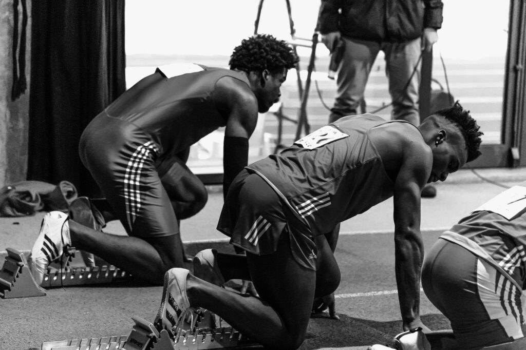 Black and white image of athletes in starting blocks ready for a sprint race indoors.