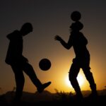 Silhouetted soccer players performing tricks at sunset, creating a scenic and dramatic view.
