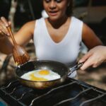 From above of crop unrecognizable multiethnic couple frying eggs on skillet using metal stove in forest camp
