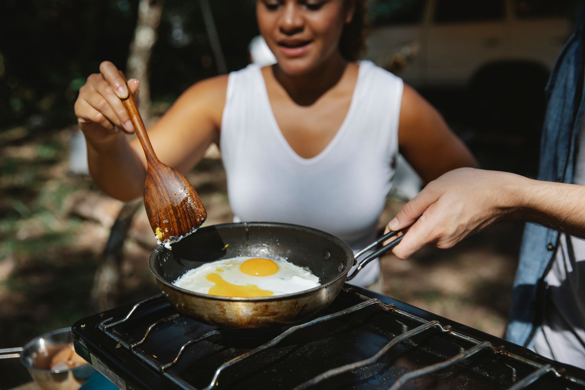From above of crop unrecognizable multiethnic couple frying eggs on skillet using metal stove in forest camp