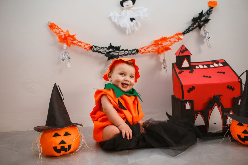 Cute baby dressed as pumpkin surrounded by Halloween decor, smiling indoors.