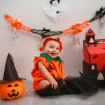 Cute baby dressed as pumpkin surrounded by Halloween decor, smiling indoors.