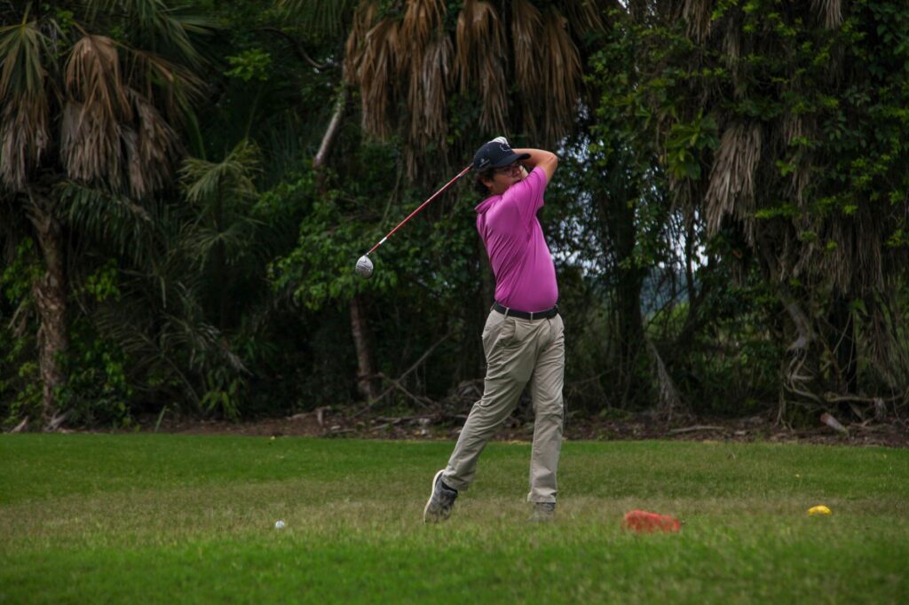 Adult male golfer swinging a club on a lush, green golf course surrounded by trees.