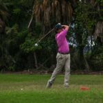 Adult male golfer swinging a club on a lush, green golf course surrounded by trees.