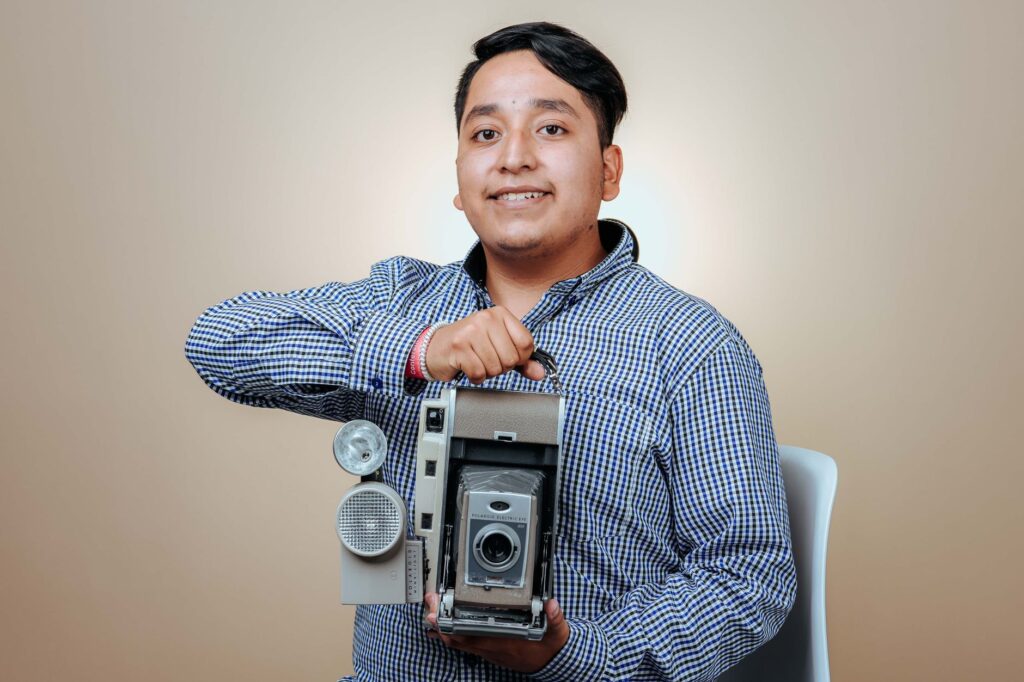 A smiling man in a checkered shirt holds a vintage camera in a studio setting.