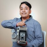 A smiling man in a checkered shirt holds a vintage camera in a studio setting.