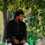 Young man in traditional attire seated outdoors, shaded by lush greenery.