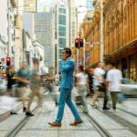 A businessman in a blue suit crosses a busy street in Sydney, Australia, surrounded by motion-blurred pedestrians.