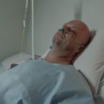 A bald man lying in a hospital bed wearing a nasal cannula for oxygen support, depicting illness and recovery.