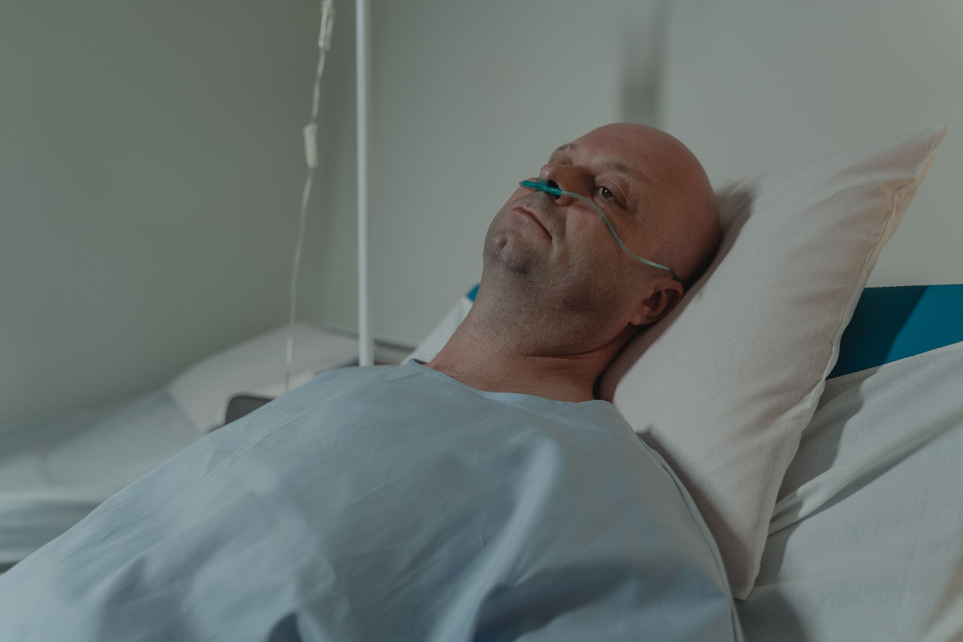 A bald man lying in a hospital bed wearing a nasal cannula for oxygen support, depicting illness and recovery.