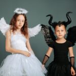 Two girls in angel and devil costumes with wings and horns against a gray background.