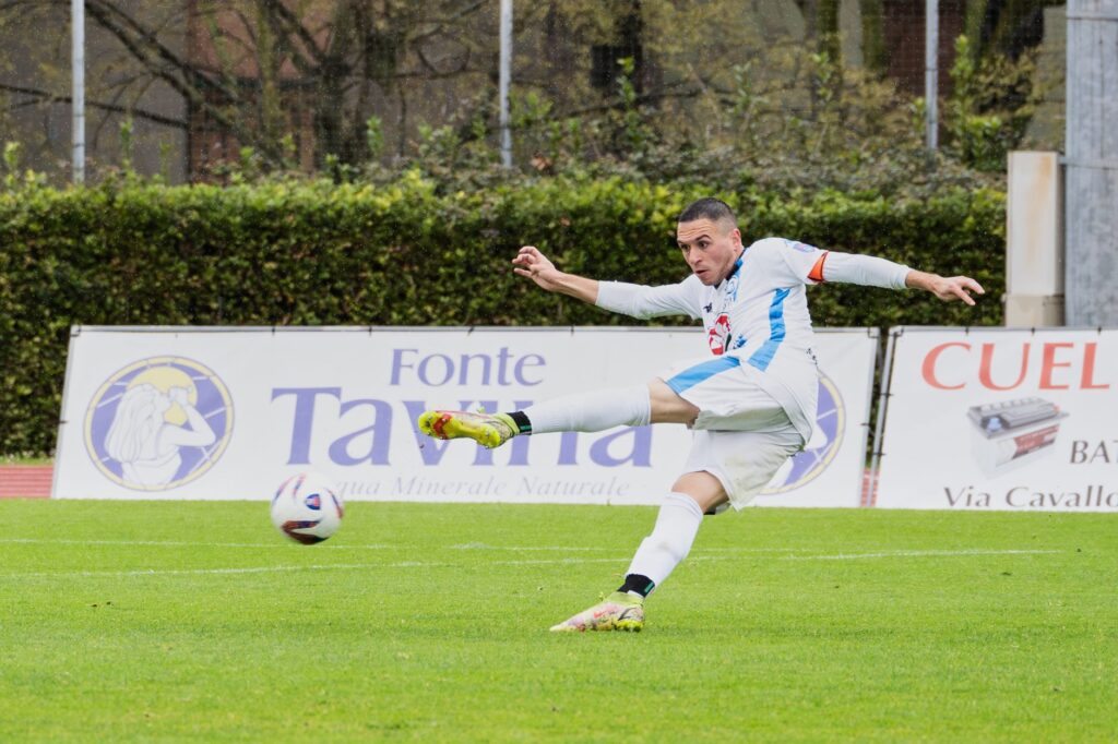 Soccer player in white kit powerfully kicking the ball during a match on a green field.