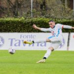 Soccer player in white kit powerfully kicking the ball during a match on a green field.