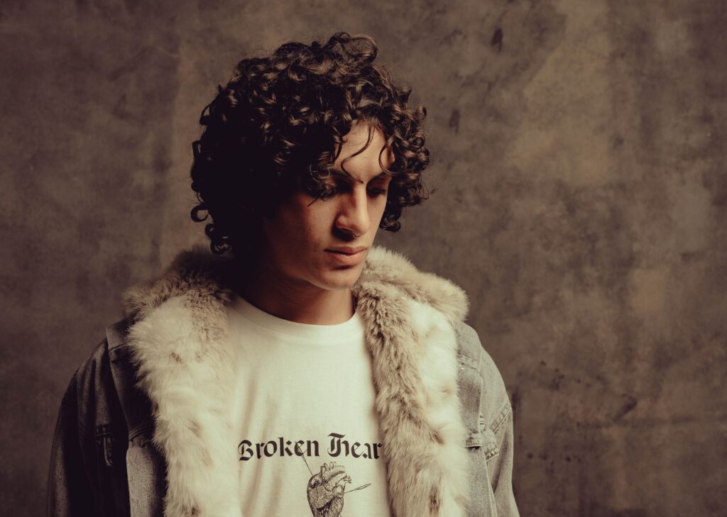 Studio portrait of a young man with curly hair wearing a fur-trimmed jacket and graphic tee.