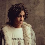 Studio portrait of a young man with curly hair wearing a fur-trimmed jacket and graphic tee.
