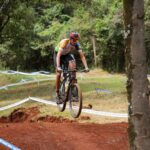 Mountain biker jumps over dirt trail in a forest competition setting.