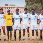 A local soccer team in uniforms posing on a grassy field under a clear sky.