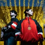 Two adults pose confidently under a canopy of glowing lights at night, wearing stylish jackets.