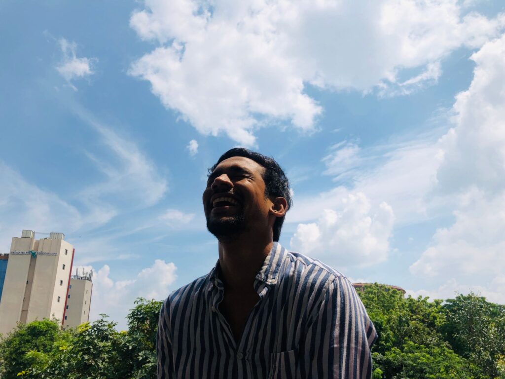 From below ethnic male in striped shirt laughing at joke against cloudy sky on sunny day in city park